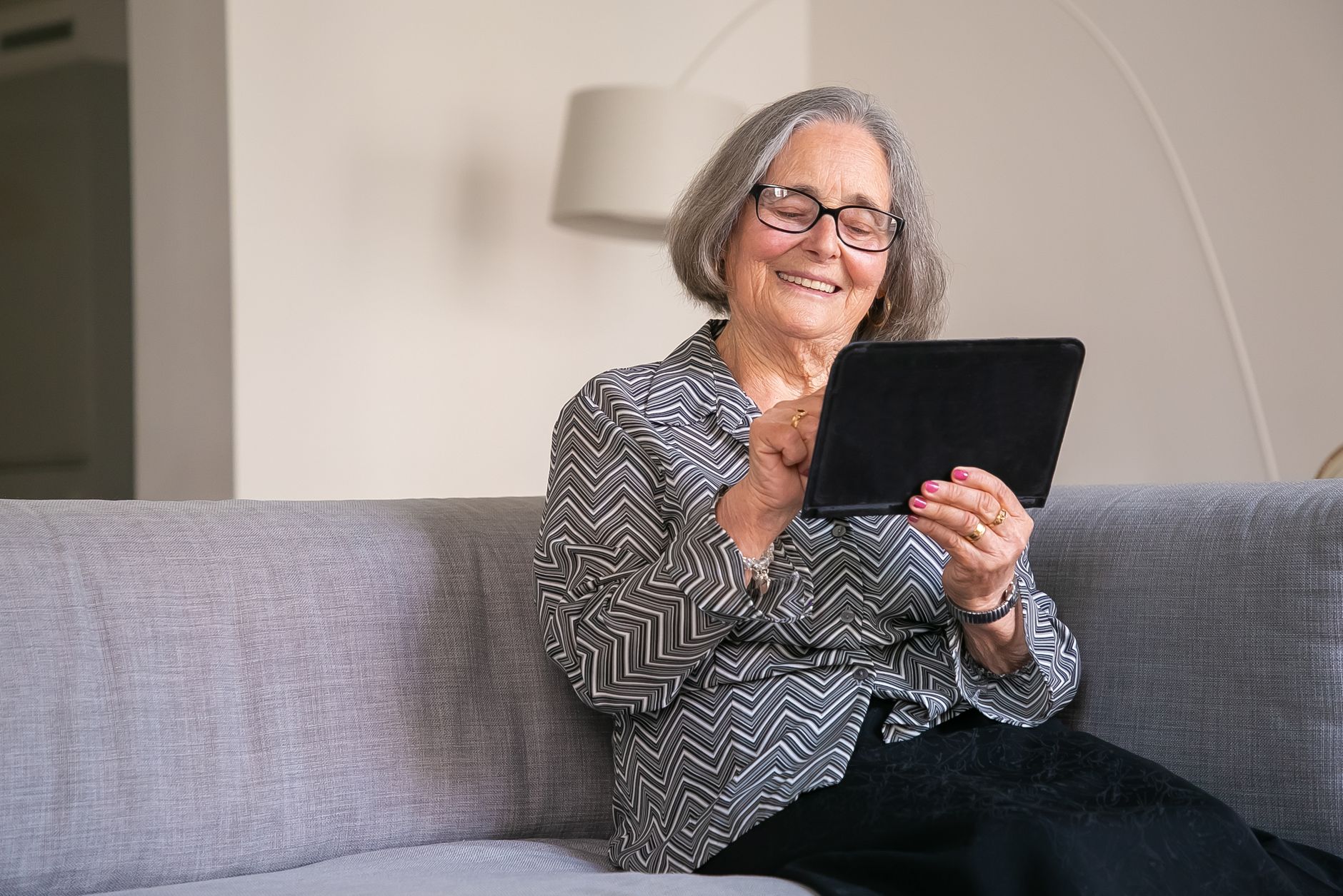 Femme souriante aux cheveux gris, installée sur un canapé clair, une tablette entre les mains