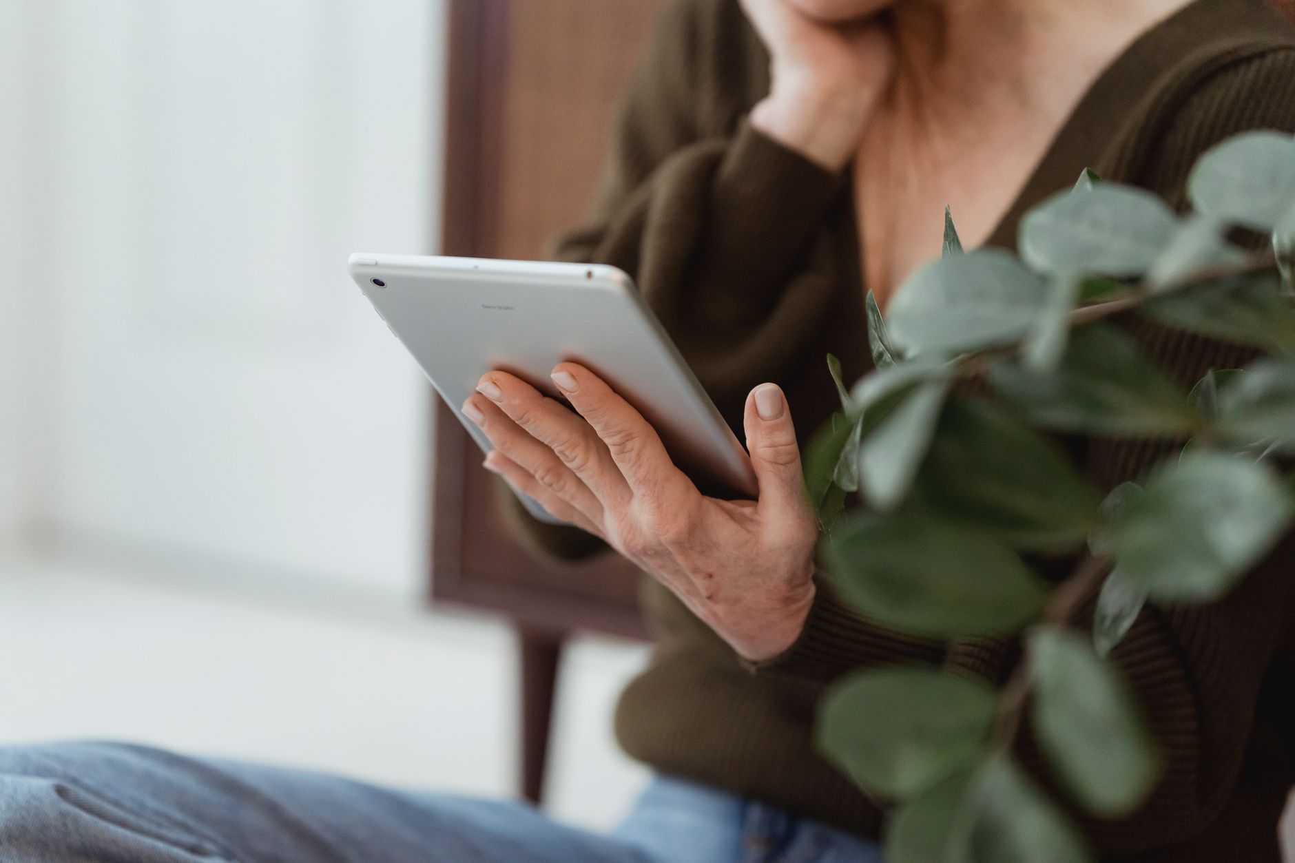 Mains d’une femme qui tient une tablette, une plante verte au premier plan, dans un salon lumineux